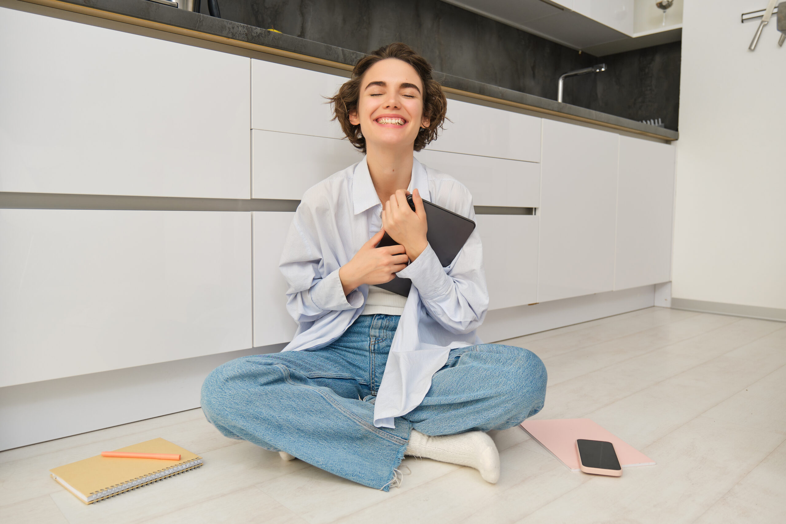 Smiling woman sitting on a kitchen floor with a notebook, surrounded by cleaning supplies.