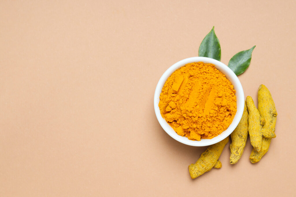Vibrant turmeric powder in a white bowl with whole turmeric roots and green leaves on a beige background.