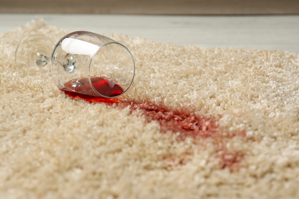 Overturned wine glass with red wine spilled on a soft beige carpet, a close-up depicting a common household accident and the need for stain removal.
